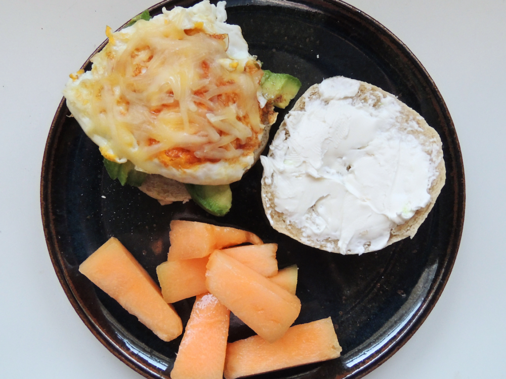 Sourdough bagel sandwich with cheese, egg, avocado, and cream cheese with a side of cantelope.