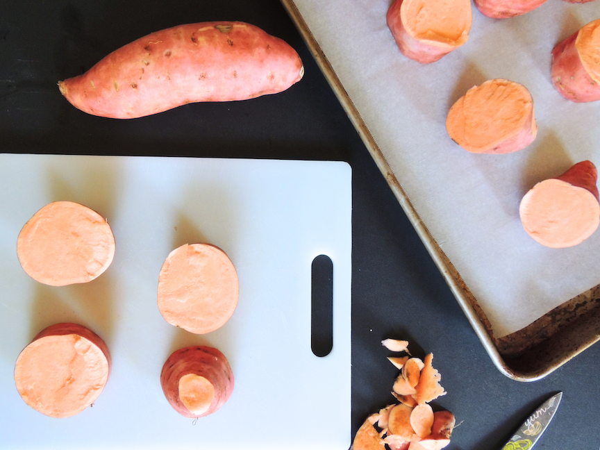 A quartered sweet potato on a cutting board, next to a baking sheet and more sliced potatoes