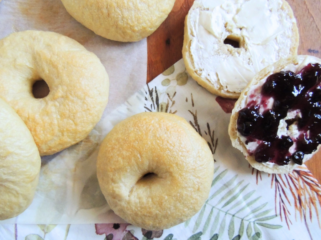 bagels sitting on a parchment paper and a napkin. One bagel is sliced and covered with cream cheese and jelly