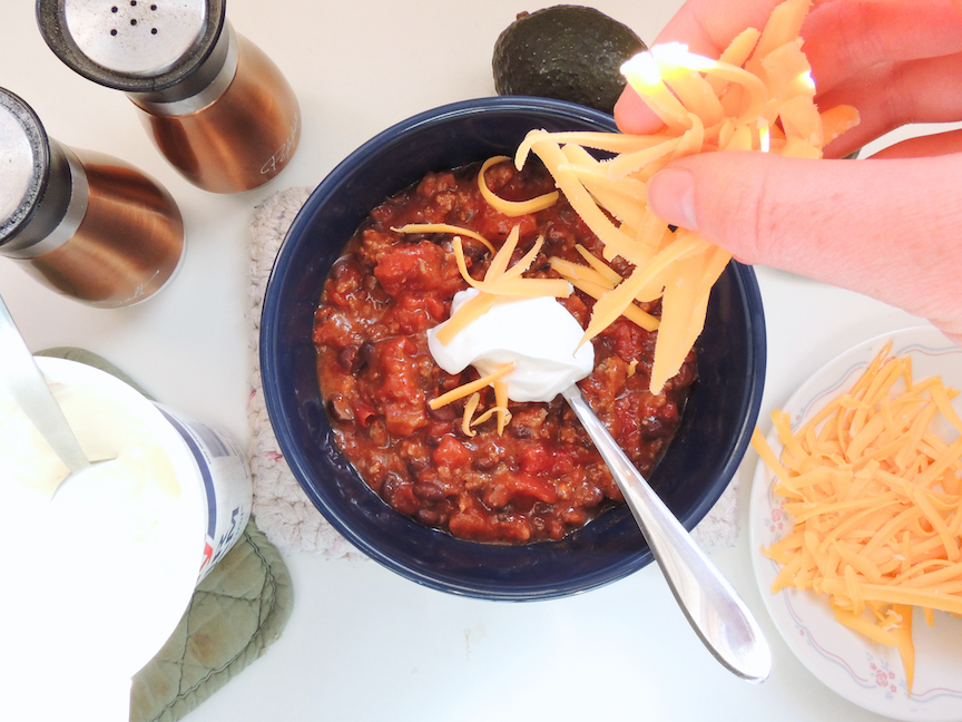 a hand sprinkling cheddar cheese over a bowl of chili